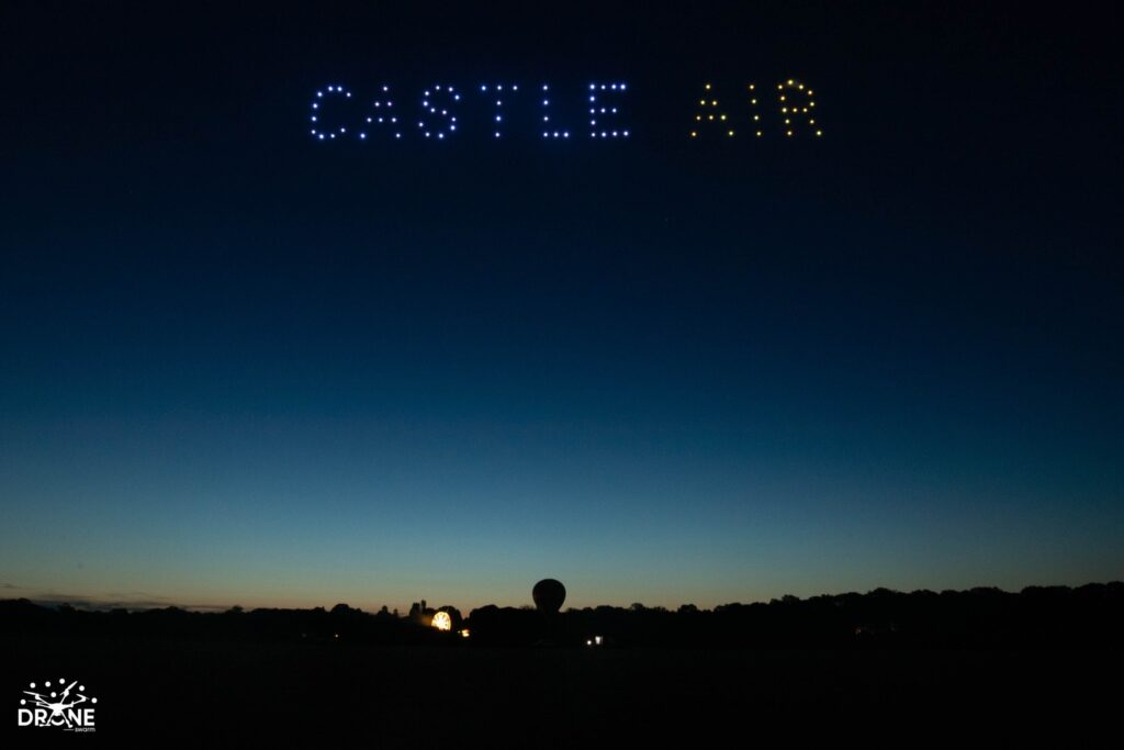 A night sky with drones forming the words "CASTLE AIR" in blue and yellow lights. Silhouettes visible below.