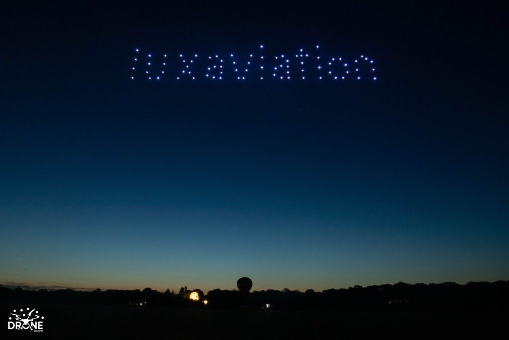 Drones light up the night sky, forming the word "Luxaviation" over a dark landscape with a hot air balloon silhouette.