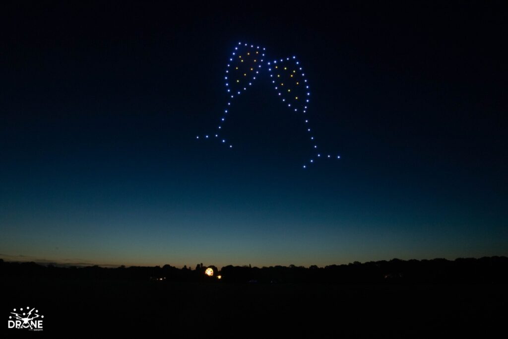 Drones forming a shape of two clinking wine glasses in the night sky with a subtle horizon line in the background.