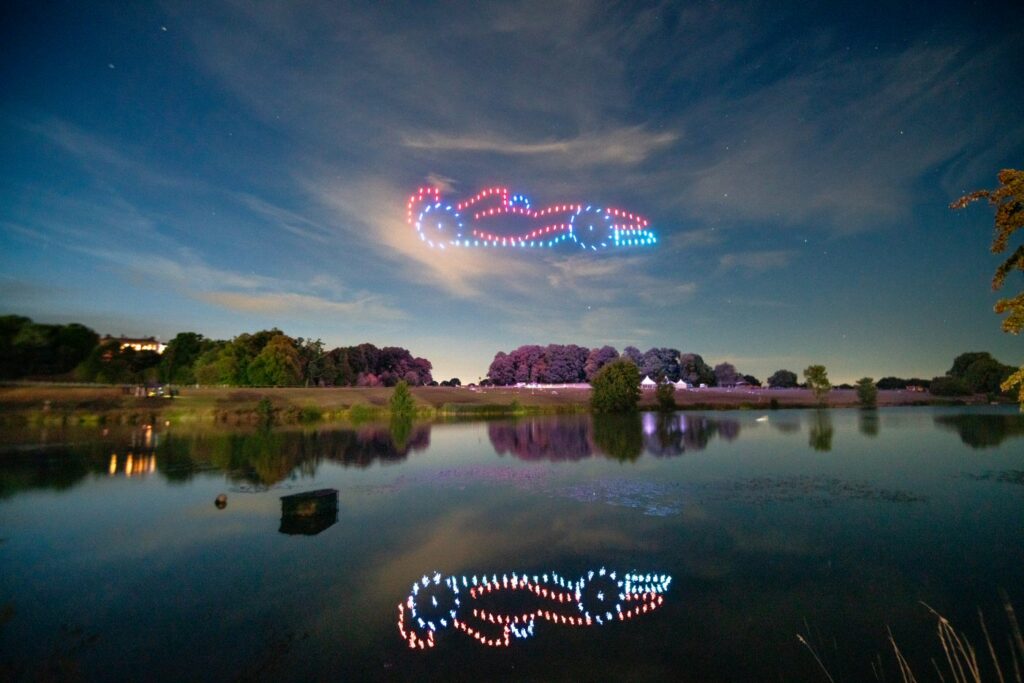 Drones form a car shape in the night sky, their reflection visible in the lake below.