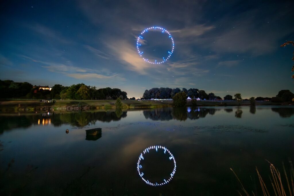 A night sky illuminated by a circular formation of lights, reflecting in a calm lake surrounded by trees.