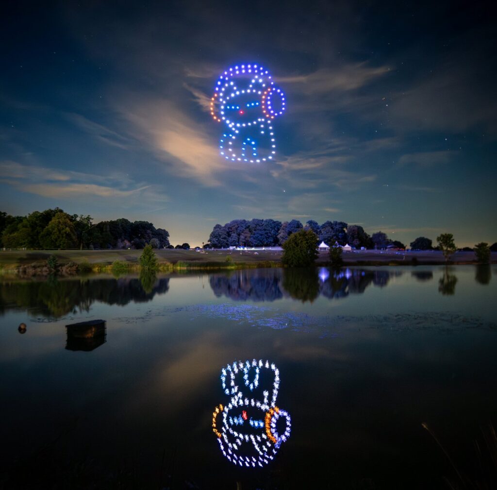 Two drone light displays of a character above a serene lake, with reflections on the water at dusk.