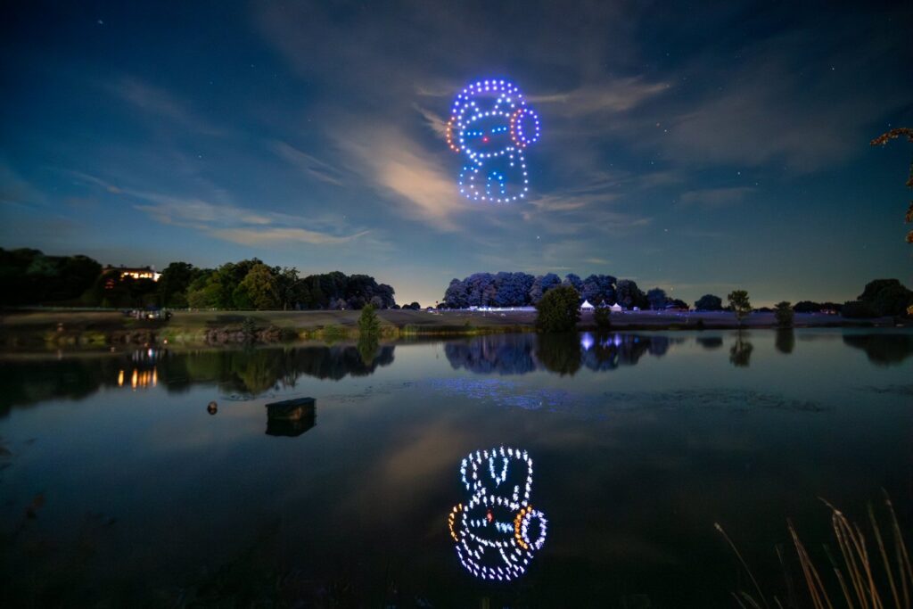 A nighttime sky with a lit-up drone formation of an animated character's face, reflected on a calm lake below.