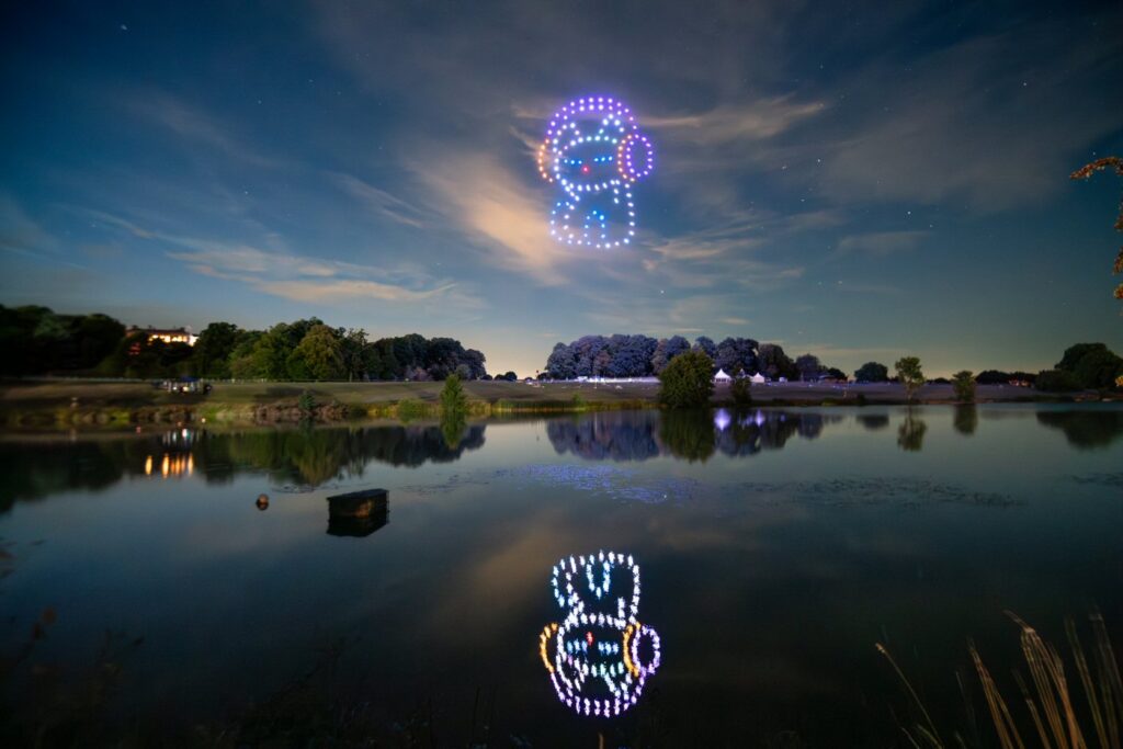 A sky is filled with drone lights in formation, creating colorful patterns that reflect on a calm lake below.