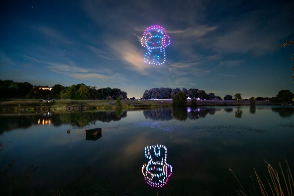 Drones form colorful Mario character in night sky, reflected in calm lake with trees and houses in the background.