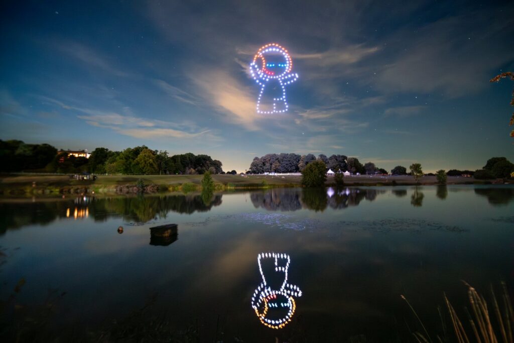 Drones in the night sky create a light display of a cartoon figure above a serene lake with trees reflecting on the water.