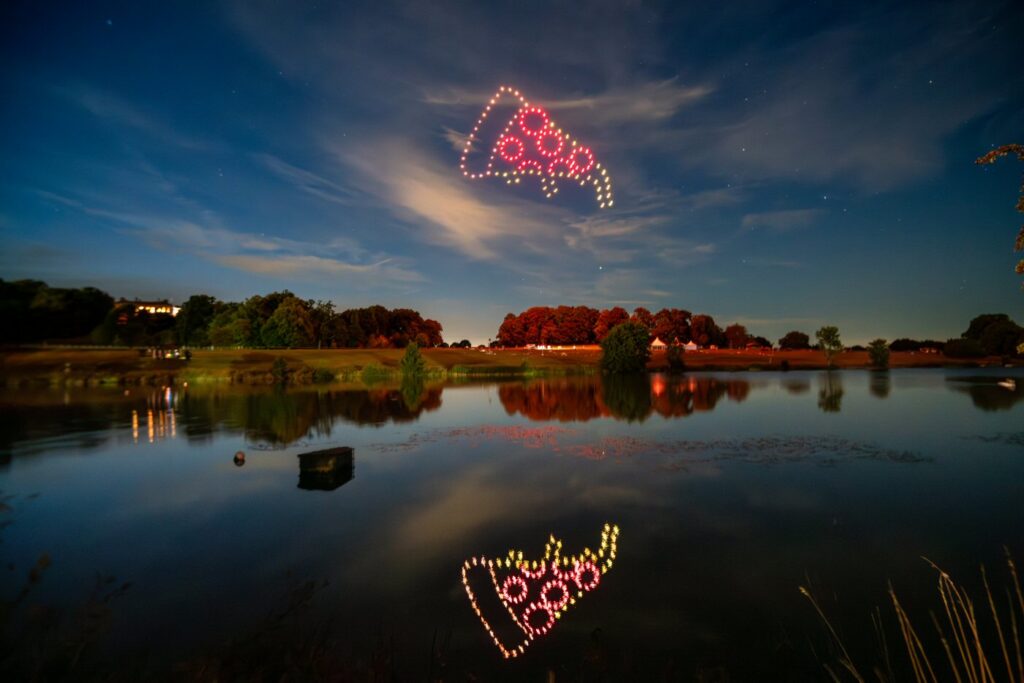 Drones form a glowing image of a pizza slice in the night sky, reflected on a calm lake surrounded by trees.