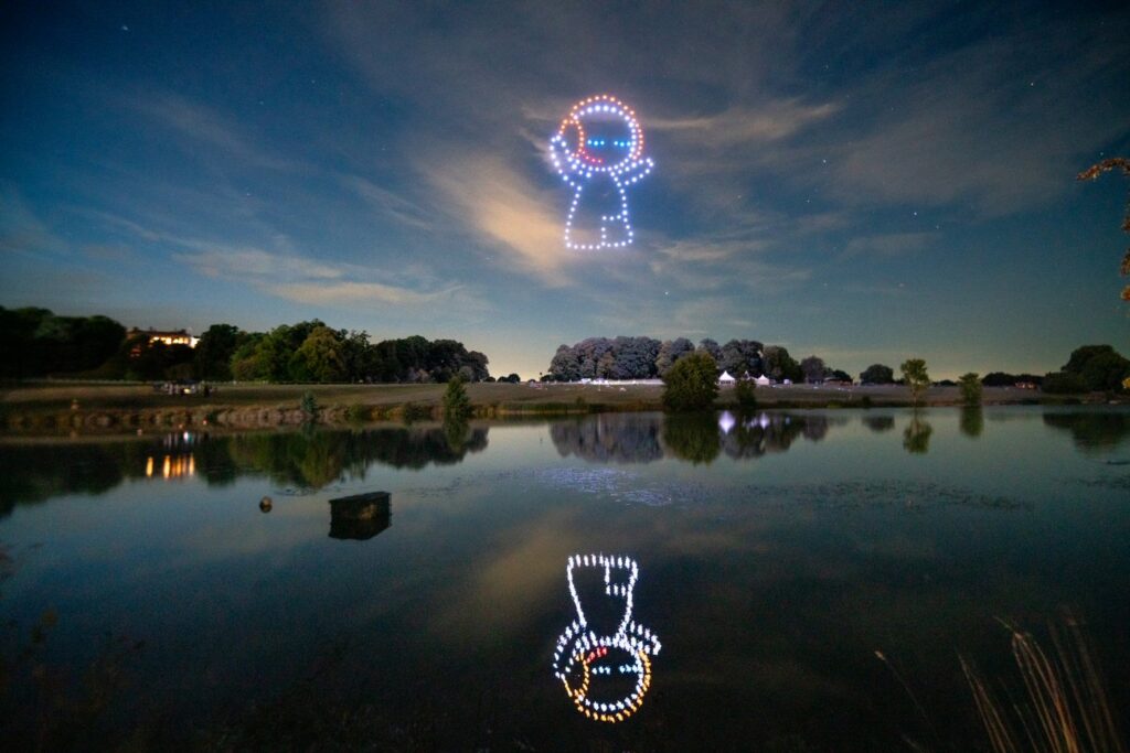 Illuminated drone display resembling cartoon characters in the night sky reflecting on a calm lake with trees in the background.