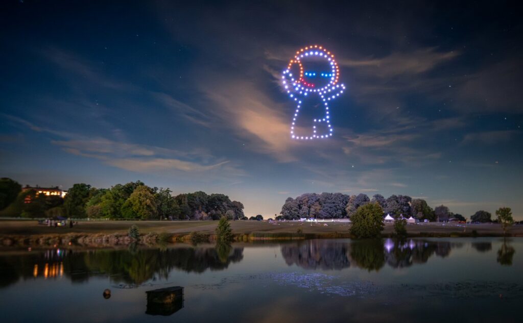 Drones form an astronaut figure in the night sky above a park with trees reflecting in a calm lake.