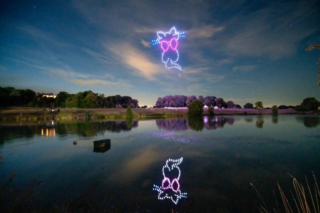 A nighttime scene with drones forming a cat wearing glasses over a reflective lake and a partly cloudy sky.