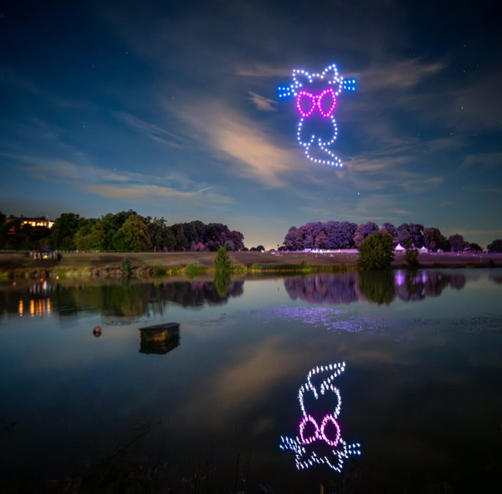 A night sky shows two owl-shaped drone displays with pink glasses, one above a lake and its reflection visible below.