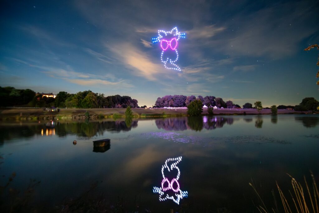 A nighttime scene with a drone light display forming a smiling face with sunglasses, reflected over a calm lake.