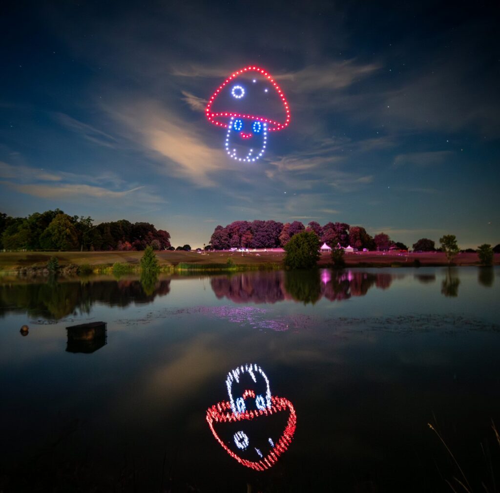 Drone show forms a mushroom and smiling face in a night sky, reflected in a tranquil lake with trees in the distance.