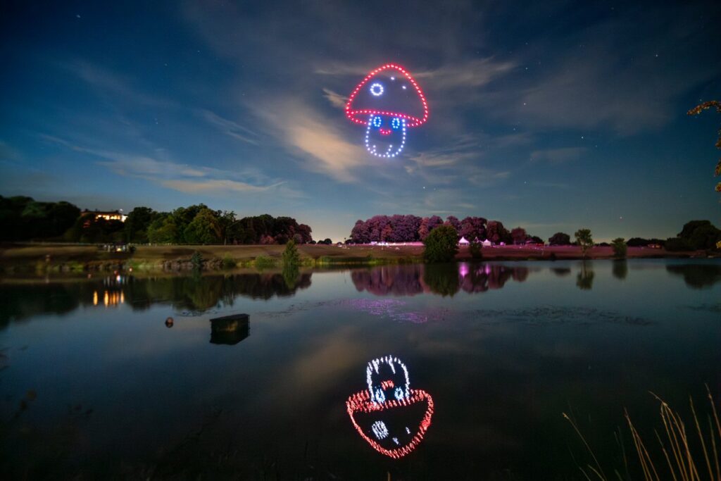 A night sky with red and blue LED drones forming a mushroom shape, reflected in a calm lake with trees in the background.