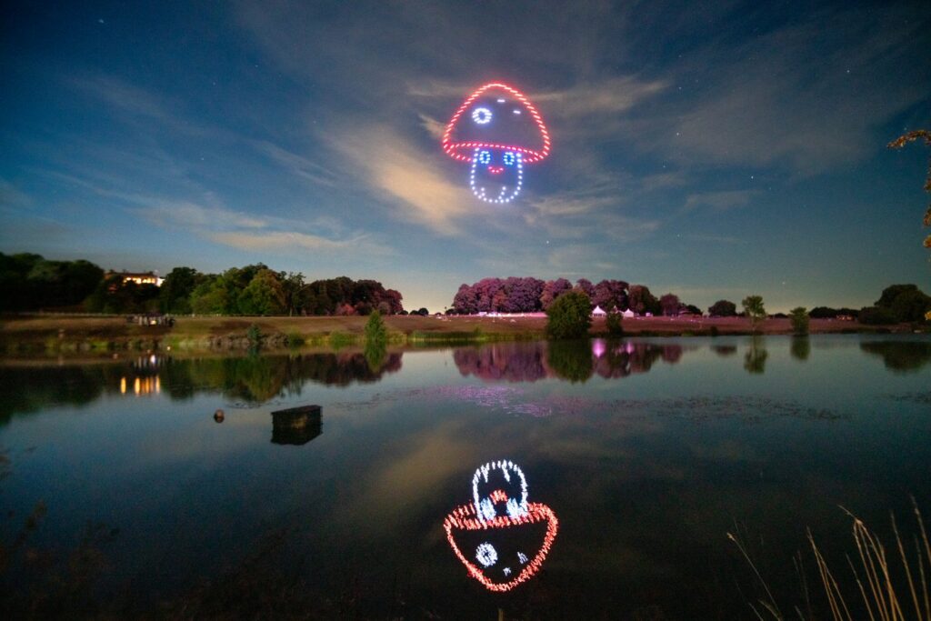 A tranquil landscape with a lake reflecting colorful mushroom-shaped drone lights in the night sky.