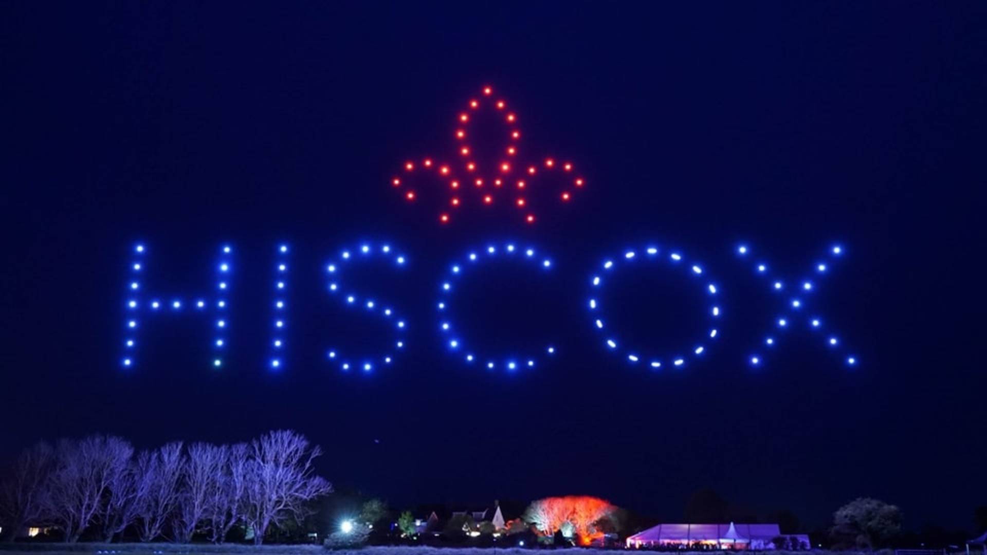 Against the night sky, drones illuminate a mesmerizing display at the Osmaston Park Wedding, showcasing "HISCOX" and a radiant fleur-de-lis.