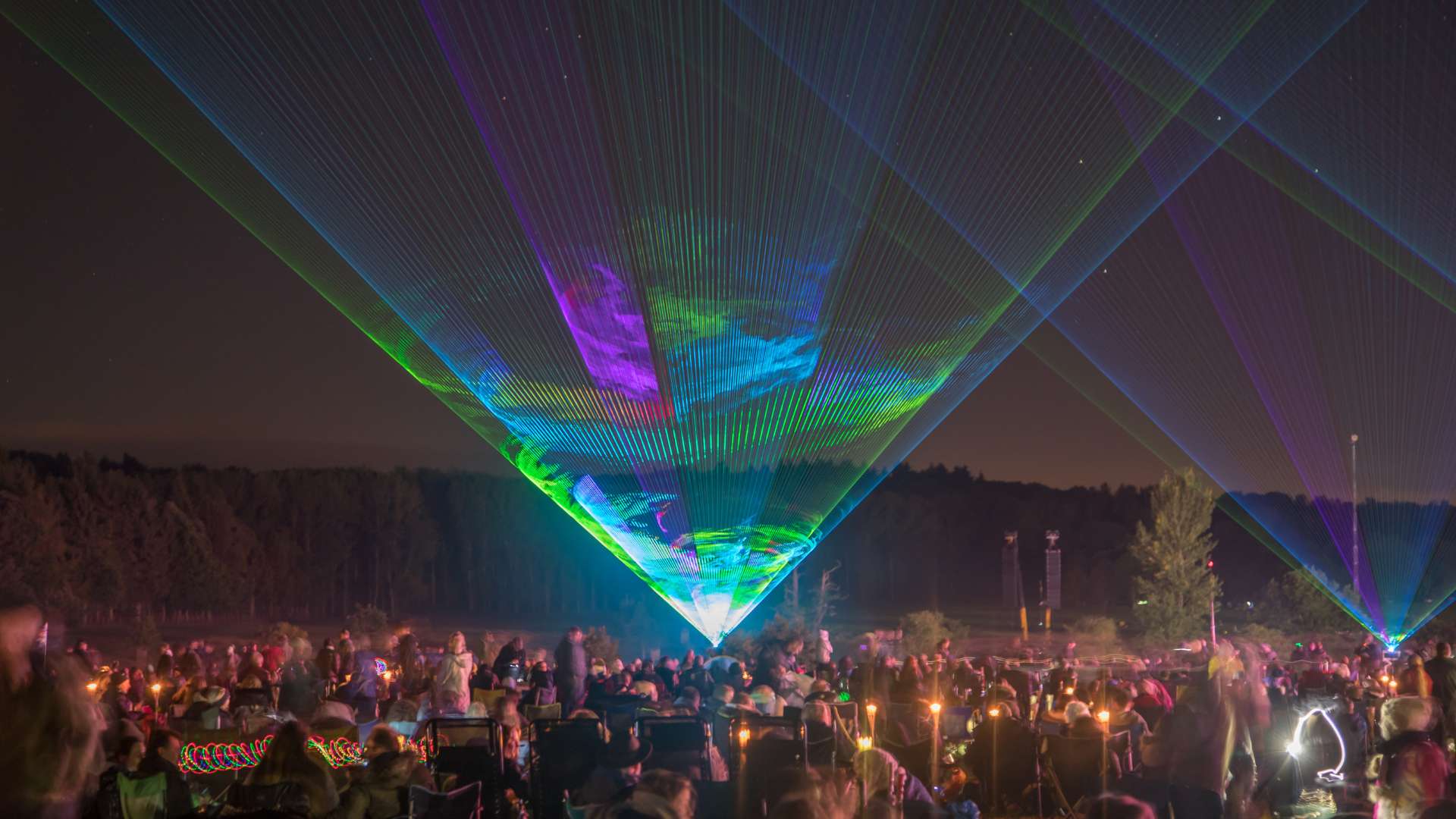 A crowd at Osmaston Park gazes in awe as a colorful laser light show fills the night sky, with green and purple beams dancing alongside a spectacular wedding drone display.