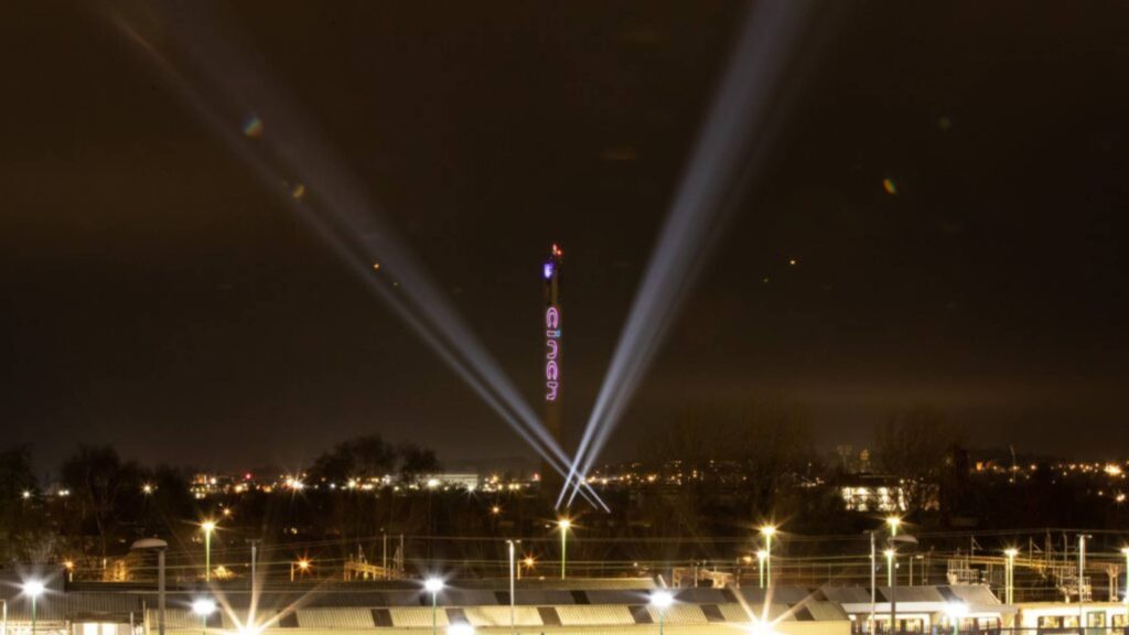 Under the city skyline at night, an illuminated sign shines brilliantly as beams of light pierce the sky, reminiscent of the dazzling displays one might expect from an Osmaston Park wedding drone show.