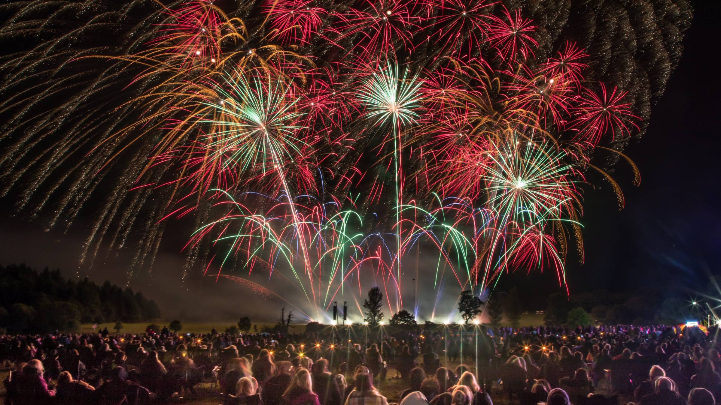 A crowd at Osmaston Park marvels as a colorful fireworks and drone show spectacularly lights up the night sky over a vast field.