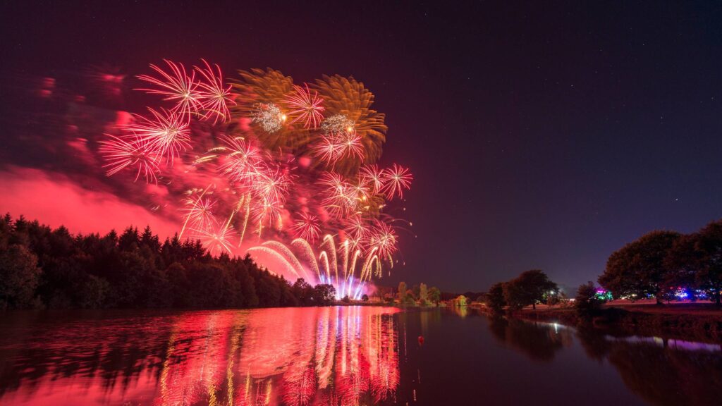 Red fireworks explode over a calm lake, their vibrant colors reflecting on the water, reminiscent of an Osmaston Park Wedding Drone Show, with trees silhouetted against the night sky.