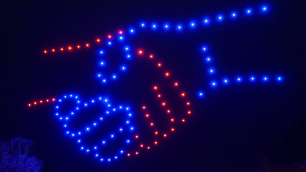 A handshake crafted from blue and red lights emerges against a dark backdrop, reminiscent of a dazzling Christmas drone light show.