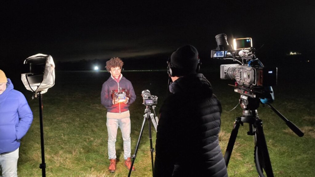 A person holding a camera lens is being filmed by a crew outdoors at night, capturing the breathtaking spectacle of a Christmas drone light show illuminating the sky.