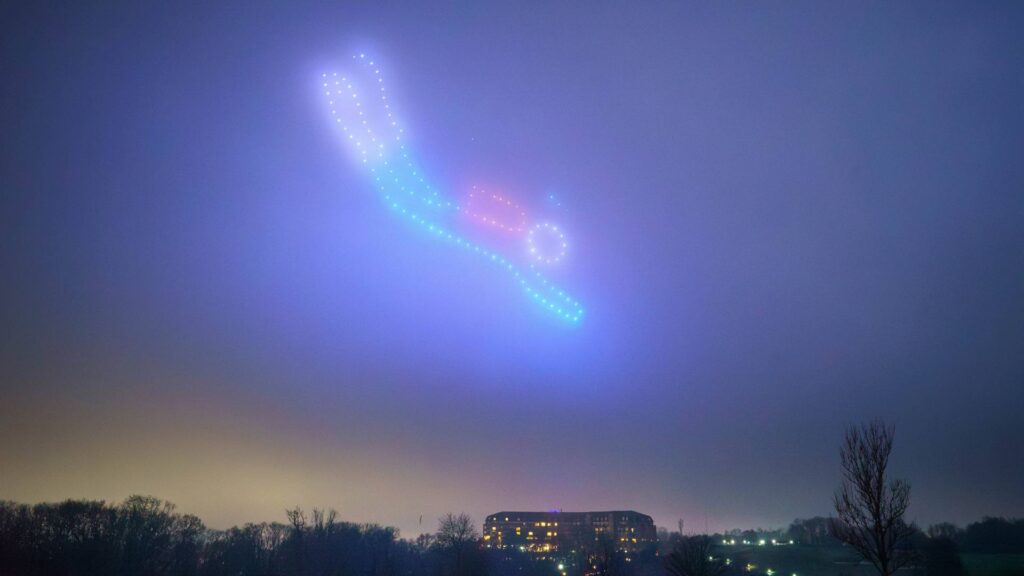 In a mesmerizing drone light show, drones form a colorful figure in the night sky above a building and trees.