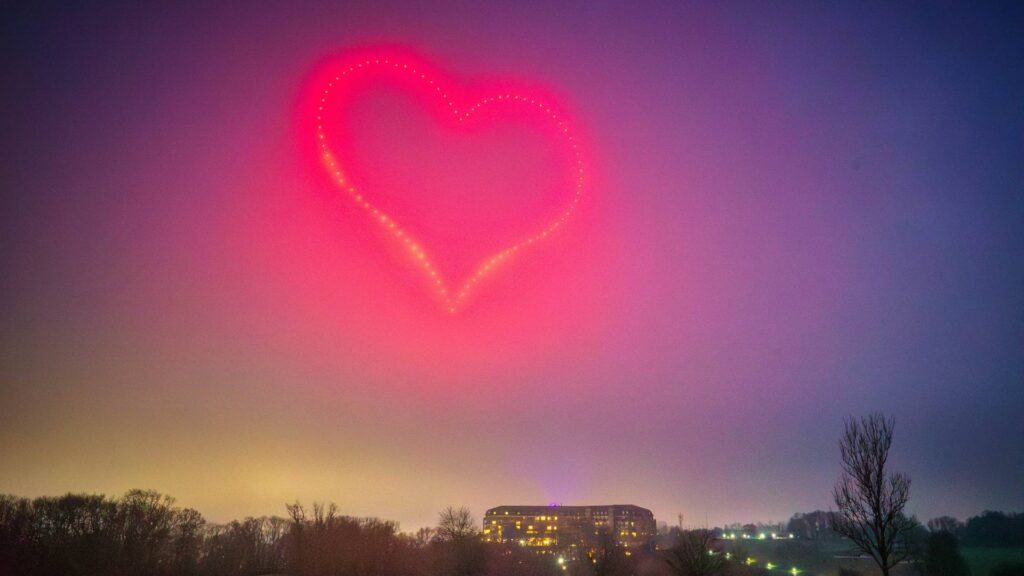 A drone light show illuminates the cloudy night sky, forming a brilliant red heart above a lit building and trees.