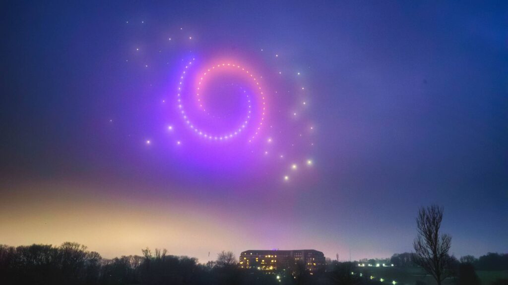 A mesmerizing drone light show spirals through the sky over a building and trees at dusk, creating a captivating display.