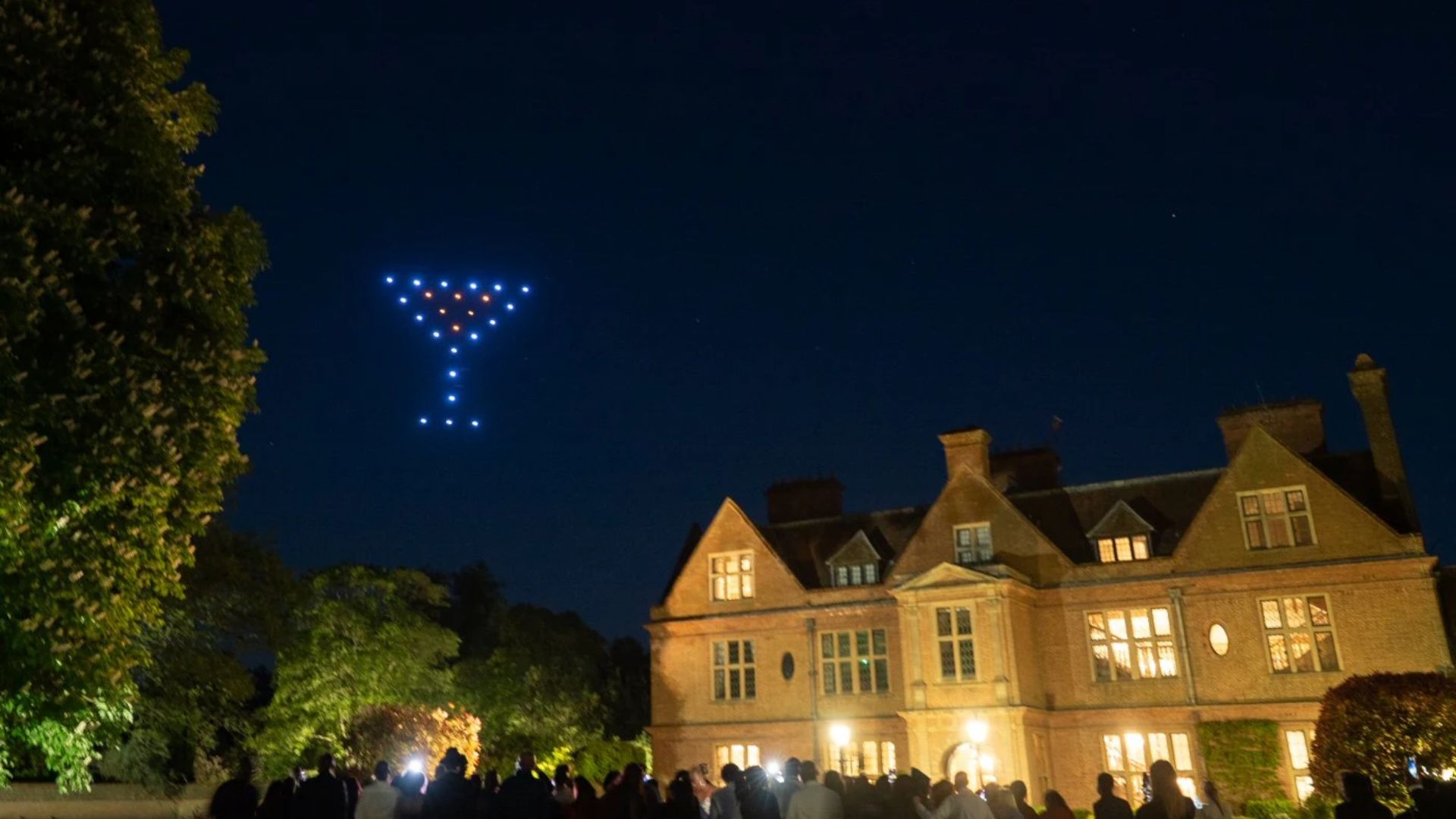 A group of drones forms a martini glass shape in the night sky above a large, lit-up building.