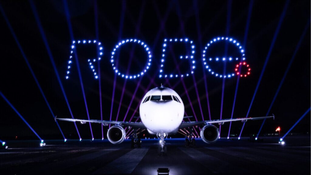 A plane on a runway at night with "ROBE" spelled out above it in blue lights from drones.