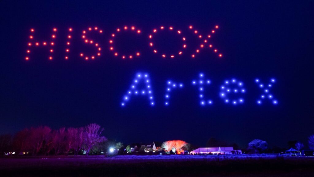 Red and blue drones spell “HISCOX Artex” in the night sky above trees and buildings.
