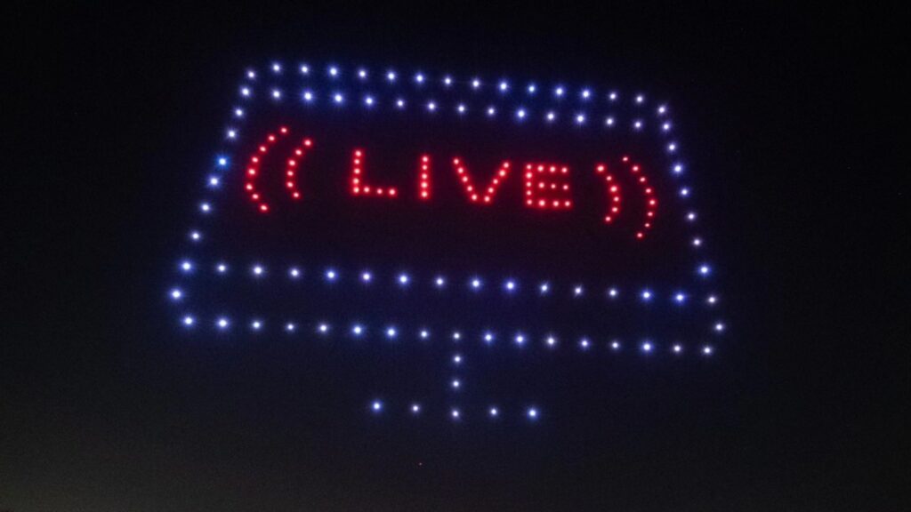 A drone light show forms a screen shape as the word "LIVE" shines in red lights against the night sky.