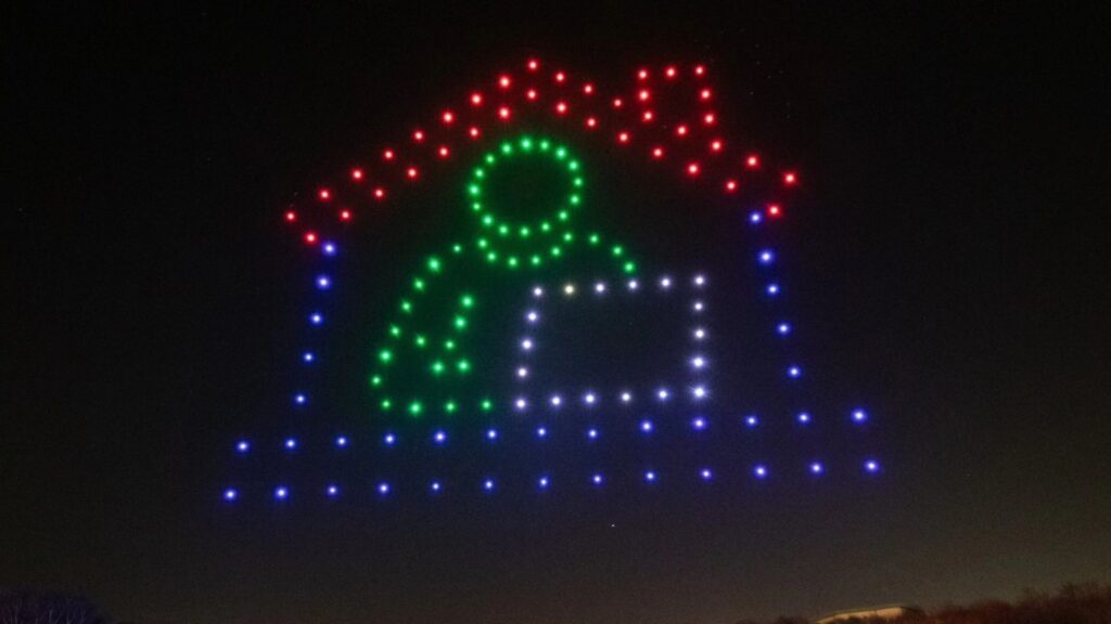 Drones form a house shape with a person inside, working at a desk, against the night sky in a stunning drone light show.