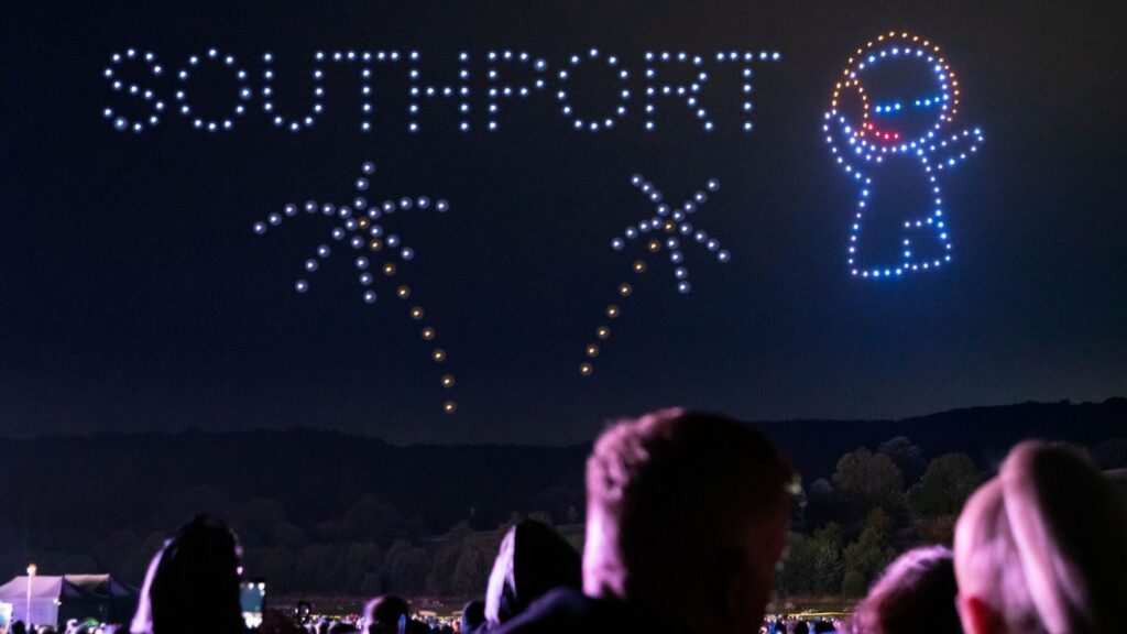 A night sky drone display spells "SOUTHPORT" with designs of fireworks and a smiling figure above a crowd.