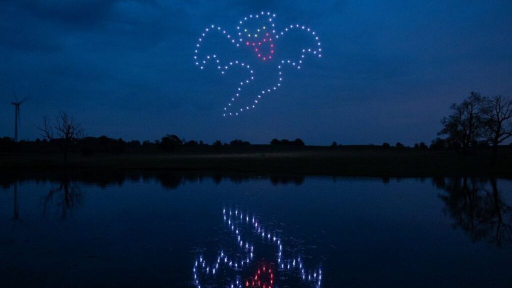 A ghost shape made of drones with lights appears in the night sky, reflecting on a calm lake below.