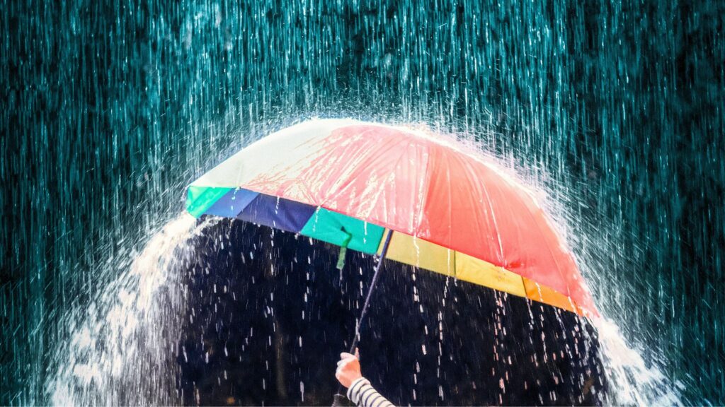 A person holds a colorful umbrella in heavy rain, with water cascading off the umbrella's edges.