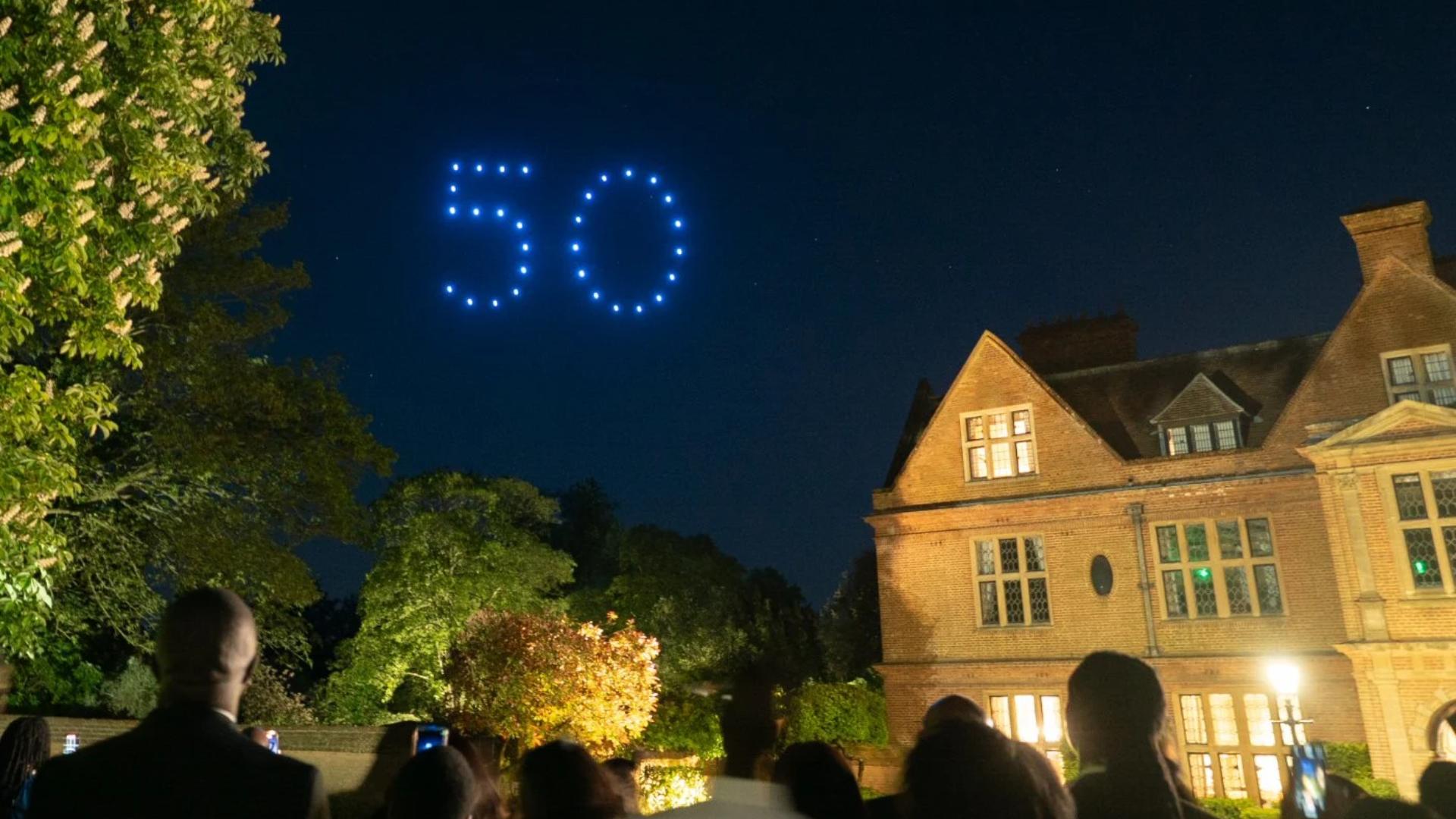 Blue drones form the number "50" in the night sky above a large brick house and trees.