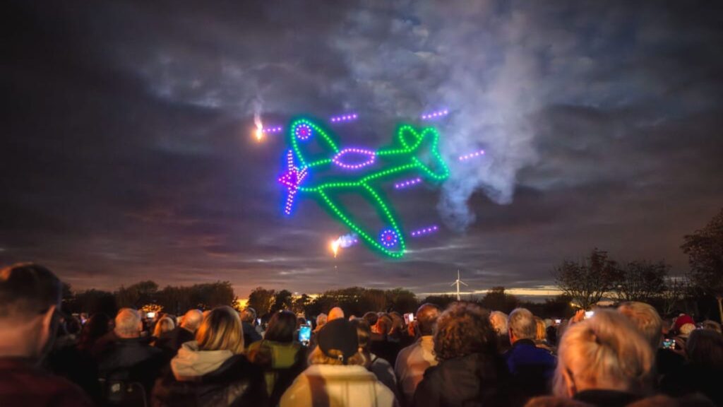 A crowd watches a colorful airplane shape formed by drones glowing in the night sky.