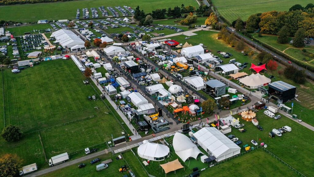 Aerial view of a large outdoor festival with white tents, booths, and parked cars on grassy fields.